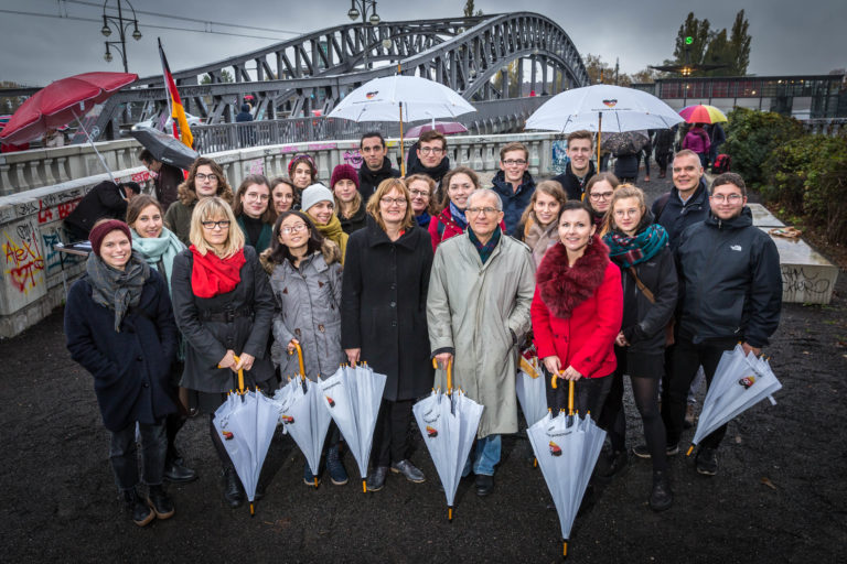Eine Gruppe von Menschen steht vor der Bösebrücke am ehemaligen Grenzübergang Bornholmer Straße in Berlin. Einige halten Regenschirme mit dem Logo des Jubiläumsjahres in der Hand.
