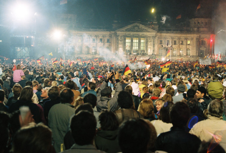 A large group of people stand on the lawn in front of the Reichstag building during a fireworks display in the night before the Day of German Unity in 1990. Some of them wave German flags.