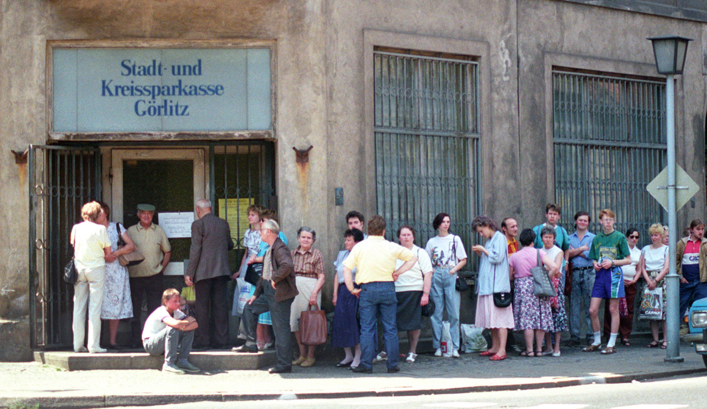 Eine lange Schlange von Menschen wartet vor einem Eckgebäude. Über dem Eingang steht ein Schild mit der Aufschrift 