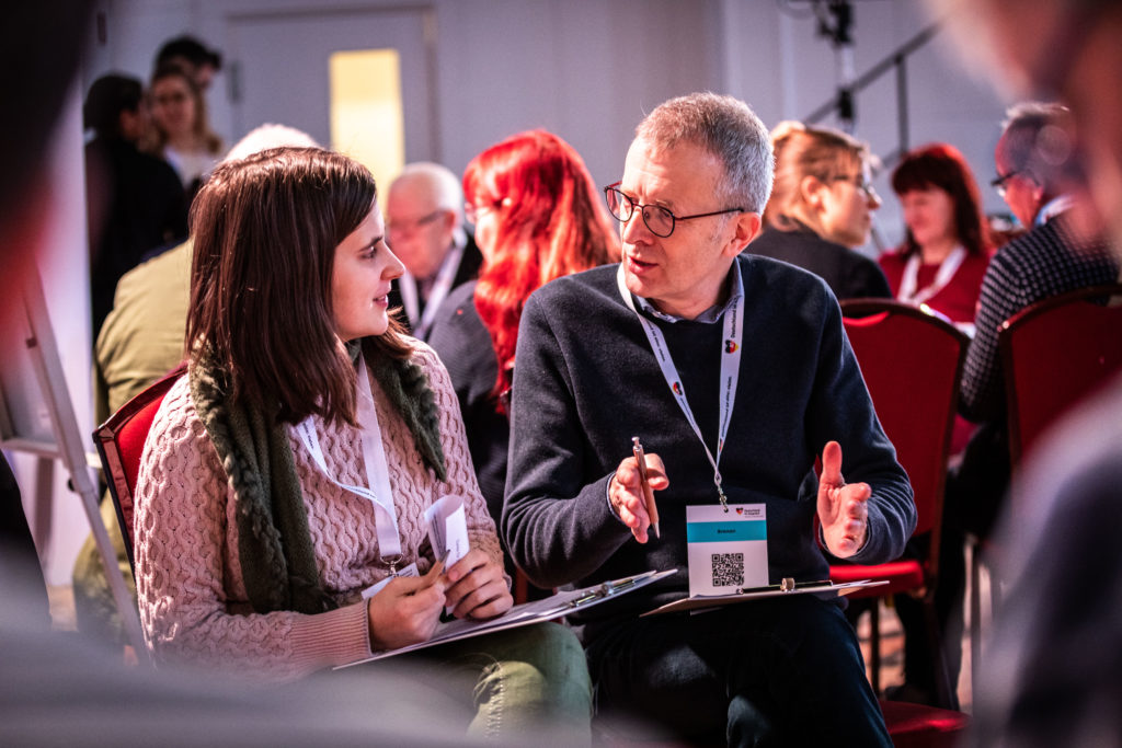 At a dialogue event in Rostock, a participant speaks with the person sitting next to him. In the background, participants are sitting at round tables holding discussions.